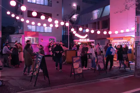 Crowds mingling under glowing pink lanterns at a Tokyo street food market at night